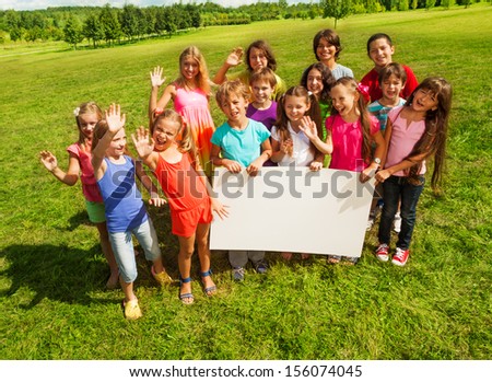 Group of kids holding blank banner showing blank placard board to write it on your own text, standing on the grass field on the park