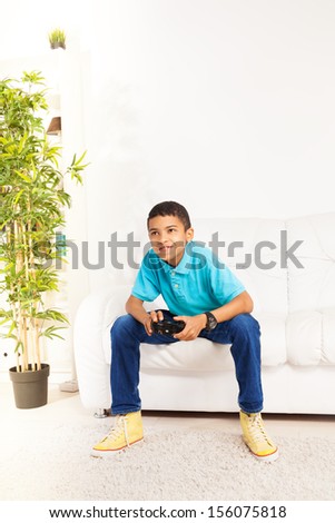 Happy black 10 years old boy playing video games holding game controller sitting on the white sofa in living room