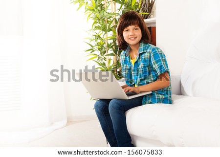 Cute little Caucasian boy with long dark hair sitting on the coach with laptop, smiling and looking at camera