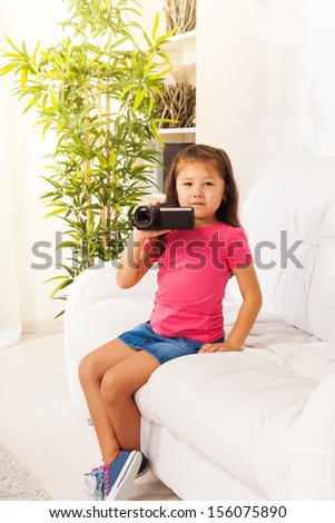 Cute little Asian girl shooting home movie with video camera sitting on the coach in living room