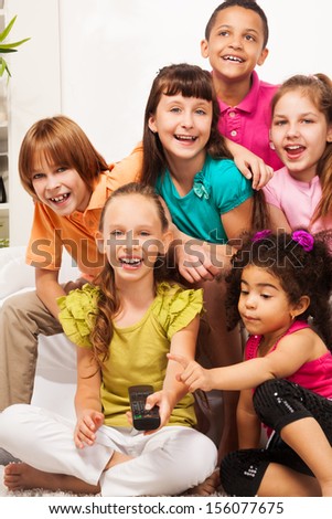 Close portrait of smiling happy girl with her friends sitting on the sofa switch on TV with remote control