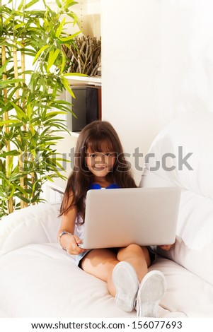 Cute girl with long dark hair sitting on the coach with laptop, smiling and looking at camera