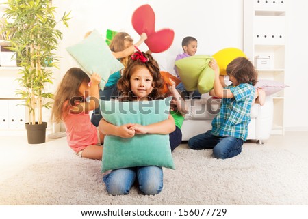 Happy smiling little Asian girl hugging pillow with large group of her friends pillow fighting on the background
