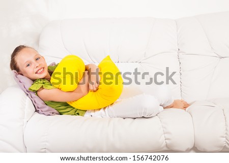Happy smiling little girl 8 years old sitting with pillow on the white leather coach in living room at home