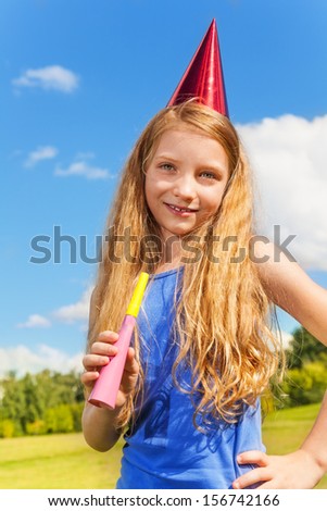 Beautiful little girl with noisemaker horn and birthday party cap standing outside with happy expression on sunny summer day