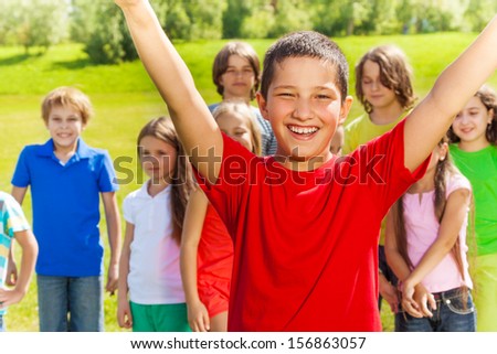 Close portrait of happy Asian boy with lifted hands and very happy expression. His friends and team standing on the background.