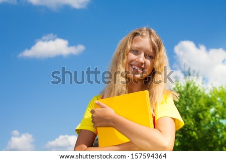 Happy blond 13 years old girl stand with with the yellow book in the park on bright sunny summer day.