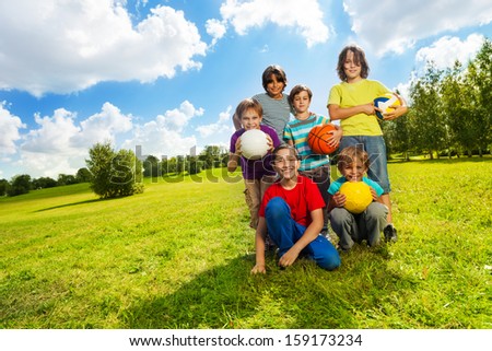 Group of very happy six boys, team sitting on the grass with balls and smiling, on sunny summer day