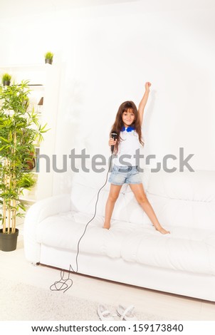 Singing cute little rock star girl with microphone and lifted hands standing on the coach in home living room