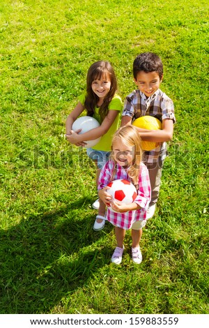 Three happy kids, boy and girls standing in the sunny summer park holding sport balls, view from top
