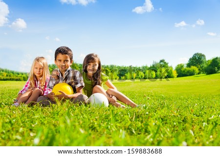 Three happy kids, boy and girls sitting in the sunny summer park holding sport balls