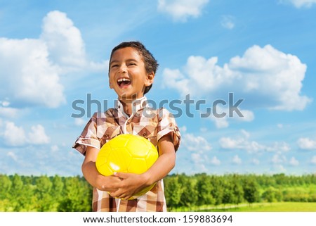 Laughing dark boy holding yellow volley ball standing in the park on sunny day with blue clouds on background