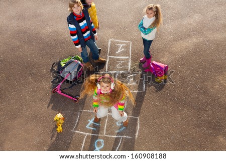 Happy girl jumping on hopscotch game with friends boys an girls standing by with school bags laying near