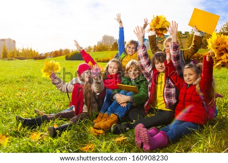 Large group of kids, boys and girls sitting in the grass in autumn clothes with maple leaves bouquets and papers after school with lifted hands after school