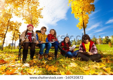 Group of five kids, friends, boys and girls, sitting on the bench in autumn park on sunny day and draw pictures