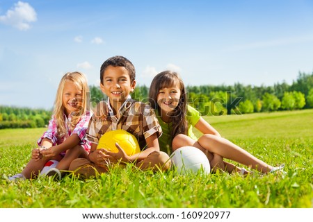 Portrait of three happy kids, boy and girls sitting in the sunny summer park holding sport balls