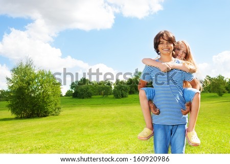 Couple school age kids girl ride on boy\'s back and hug guy, happy standing outside in the park on sunny summer day