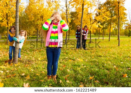 Group of five kids play hide and seek with girl counting and friends hiding behind the trees