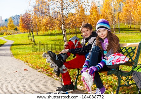 Children putting on roller blades 10 and 11 years old couple of school kids, boy an girl in warm autumn clothes on the bench in park