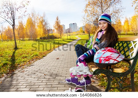11 years old beautiful girl sitting on the bench in the park putting on roller blades to go skating in the autumn park on sunny day