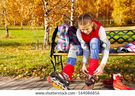 Happy blond 11 years old girl with amazing smile putting on roller blades sitting on the bench in the autumn park on sunny day, front view
