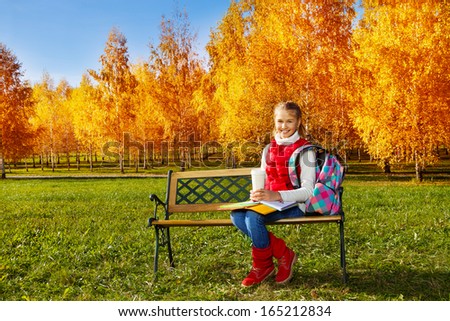 Happy blond 11 years old girl with amazing smile with books, textbooks, coffee and backpack sitting on the bench in the autumn park on sunny day