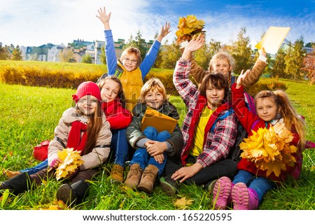 Large group of kids, friends, boys and girls around 10 years old, sitting in the grass in autumn clothes with maple leaves bouquets and papers after school with lifted hands