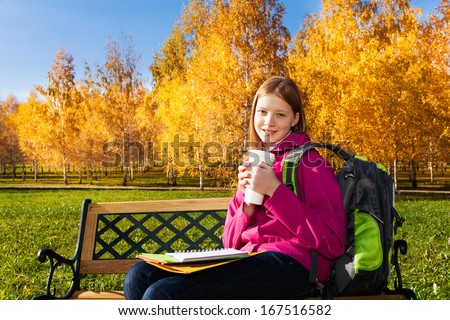 Beautiful teen 14 years old school girl sitting on the bench in autumn park wearing backpack with coffee