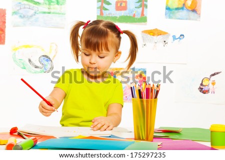 Cute little 3 years old girl in yellow shirt and pony tails draw with pencil in the art class with images on the wall