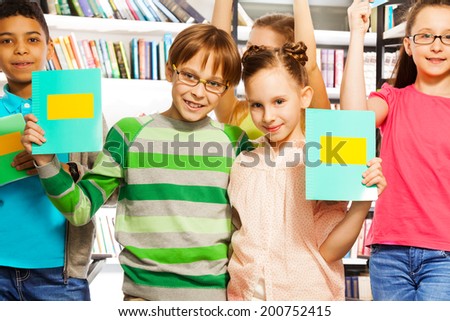 Boy and girl hold exercise books in library with three other friends behind