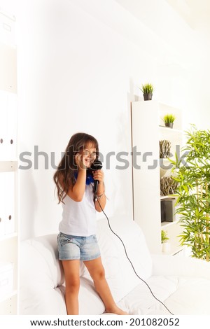 Singing cute little happy girl singing to microphone s both standing on the coach in home living room