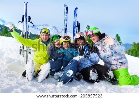 Smiling friends after skiing sitting on snow hug and wave hands with ski standing behind