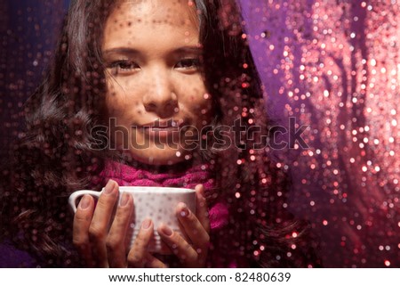 Positive Asian woman behind the glass in rainy weather with cup of hot tea