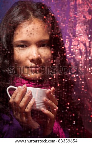 Positive Asian woman behind the glass in cold rainy weather with cup of hot tea