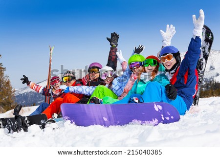 Seven happy smiling friends wearing ski mask sitting together lifting hands up in the air on the mountains background
