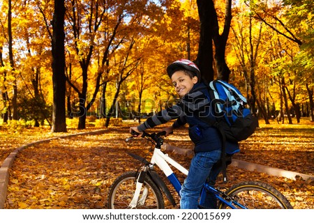 Happy smiling 8 years old black boy with backpack riding a bike in the autumn park full of orange leaves leaning on bicycle stern turning back