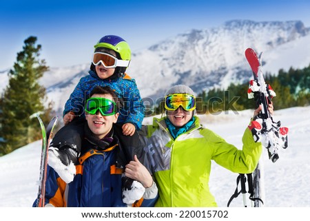 Mother, father with child on his shoulders in ski masks holding ski on mountains background