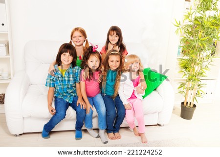 Group of 6 happy little kids sitting together on the couch at home interior in living room smiling and having fun a