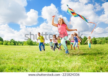 Happy little girl running with kite and her friends on the summer green field on sunny day