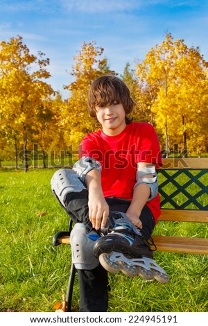 Happy 12 years old boy in red shirt sitting on bench and putting on skates in the autumn park