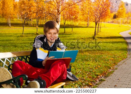 10 years old boy sitting on the bench in autumn park with smile on the on his face and reading from textbook