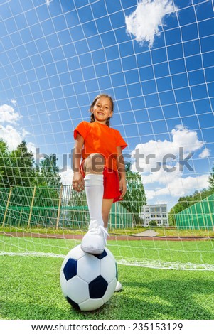 Girl with leg on football stands in front of white net of woodwork on sky background