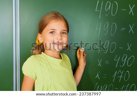 Cute smiling girl with chalk in her hand near  mathematics equation looking straight in front of blackboard during mathematics lesson
