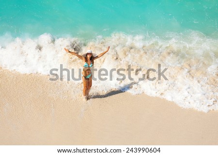 View from top of girl with white hat in striped bikini standing in ocean waves on the sand of beach during holidays summer time