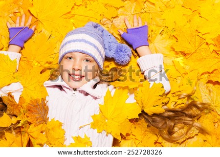 Happy school age Caucasian girl laying on the ground covered with autumn leaves, smiling and with lifted hands