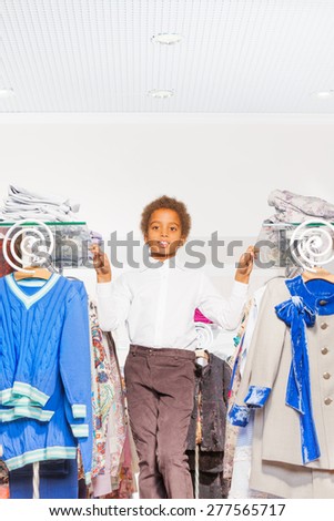 Smiling African boy between rows with clothes on the hangers and glass shelves with piles of clothes in the shopping mall