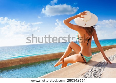 Beautiful back of young woman with white hat sunbathes under sun while sitting on stoned pier near to the ocean in summer