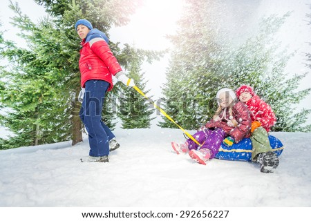 Children sitting on snow tube and other boy pulling them in winter during day in the fir tree forest