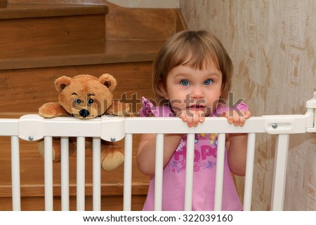 Little blonde girl on the stairs with a gate with a teddy bear friend