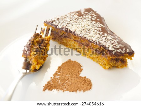 A piece of carrot cake with chocolate cream and sesame, a cinnamon heart, a fork, on a white plate